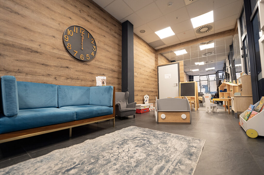 Ein modernes Wartezimmer mit blauer Couch und Spielbereich für Kinder, Holzwände und großer Uhr.