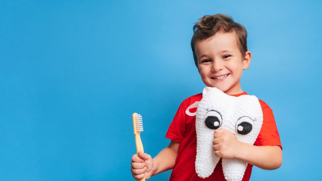 Ein lächelnder Junge mit gesunden Zähnen hält einen Plüschzahn und eine Zahnbürste auf einem blauen, isolierten Hintergrund. Mundhygiene. Pädiatrische Zahnmedizin. Prävention von Karies. Ein Platz für Ihren Text.