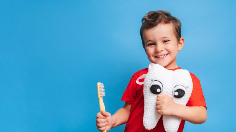 Ein lächelnder Junge mit gesunden Zähnen hält einen Plüschzahn und eine Zahnbürste auf einem blauen, isolierten Hintergrund. Mundhygiene. Pädiatrische Zahnmedizin. Prävention von Karies. Ein Platz für Ihren Text.